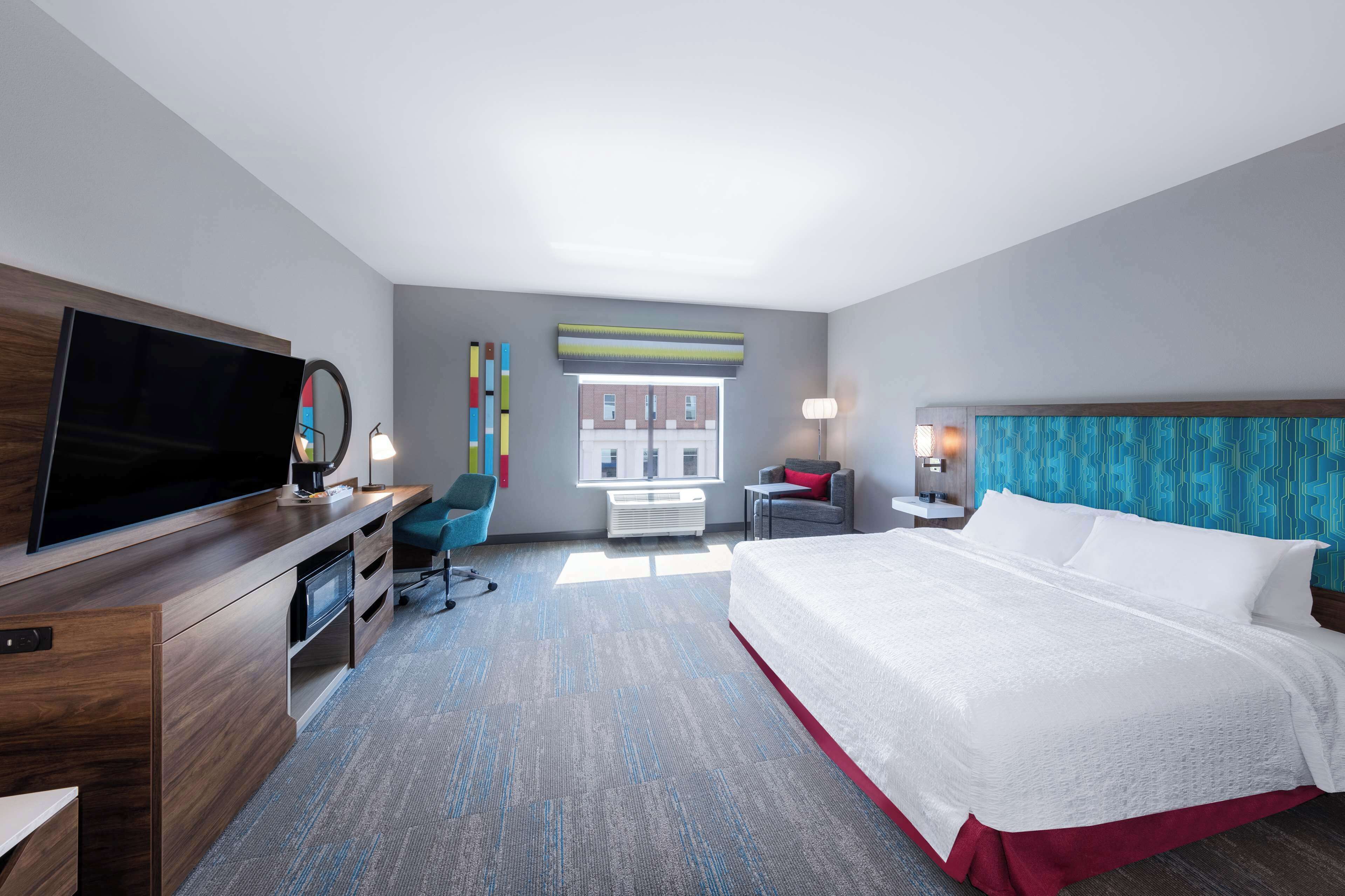 A hotel room with a large bed, a desk with a flat-screen TV, a chair, a desk lamp, and a window with blinds.