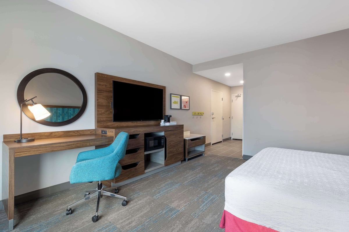 A hotel room with a wooden desk, a blue chair, a bed, a television, and a microwave.