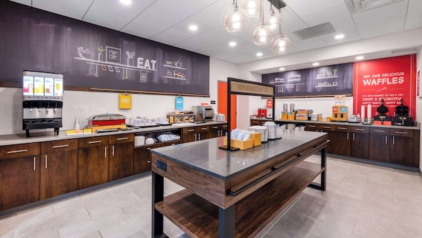 A modern cafe with a central wooden table, a coffee machine, and a waffle station. - Hampton Inn Columbus Downtown, GA (Columbus)