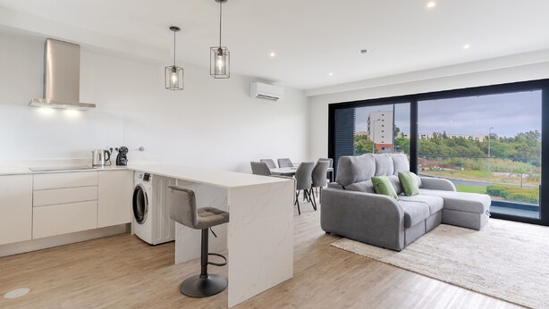 A modern kitchen with a built-in washing machine, a bar area, and a grey sofa in a living area with a large window. - Apartamento Orquídea (Funchal)