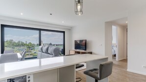 A modern living room with a large window, a flat-screen TV, a dining area, and a small kitchen. - Apartamento Orquídea (Funchal)