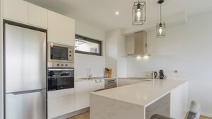 A modern kitchen with white cabinetry, a built-in oven, a microwave, and a large island with a marble countertop. - Apartamento Orquídea (Funchal)