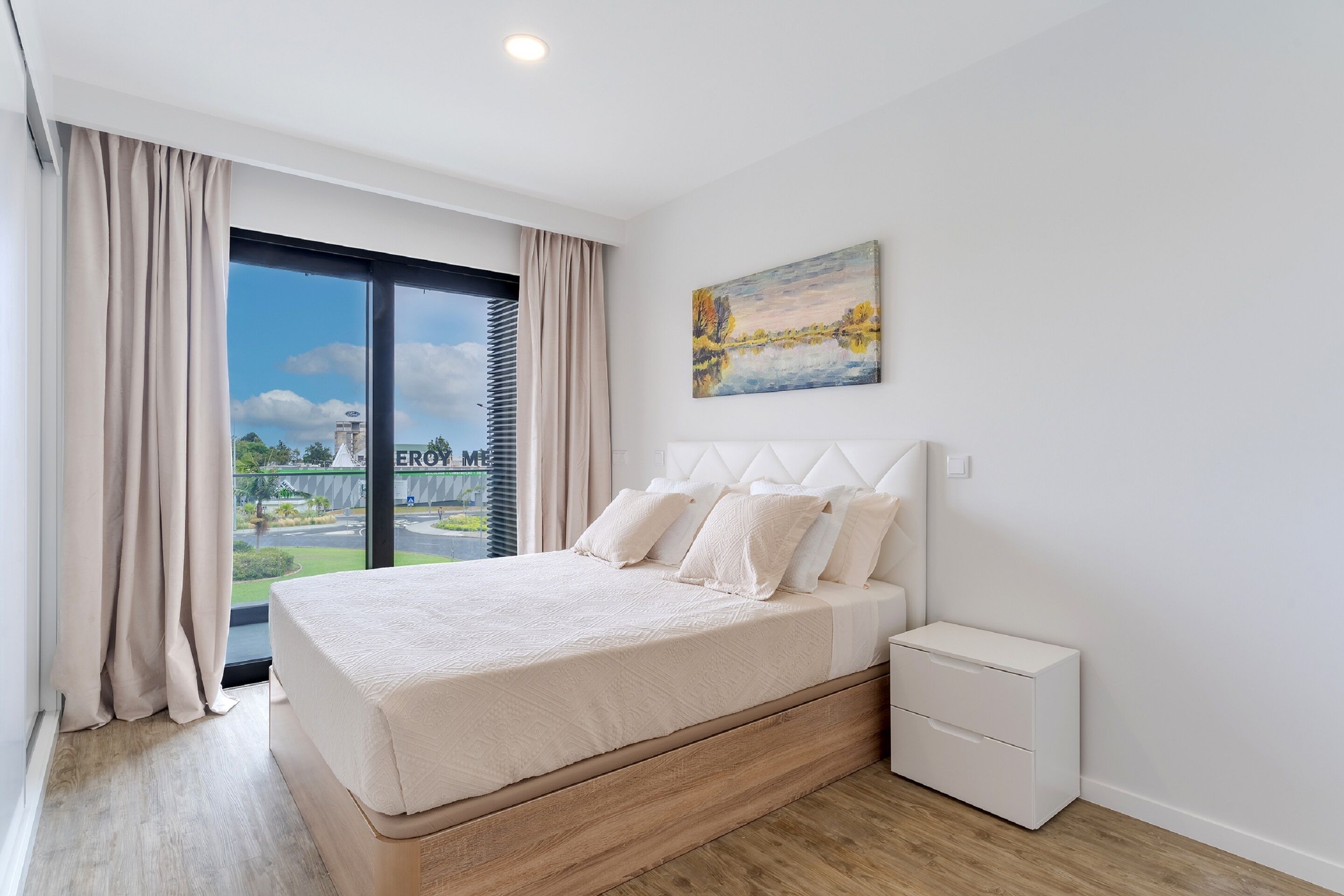 A modern bedroom with a large bed, a nightstand, a painting, and a view of a building and greenery outside.