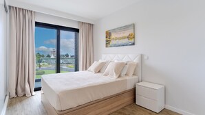 A modern bedroom with a large bed, a nightstand, a painting, and a view of a building and greenery outside. - Apartamento Orquídea (Funchal)