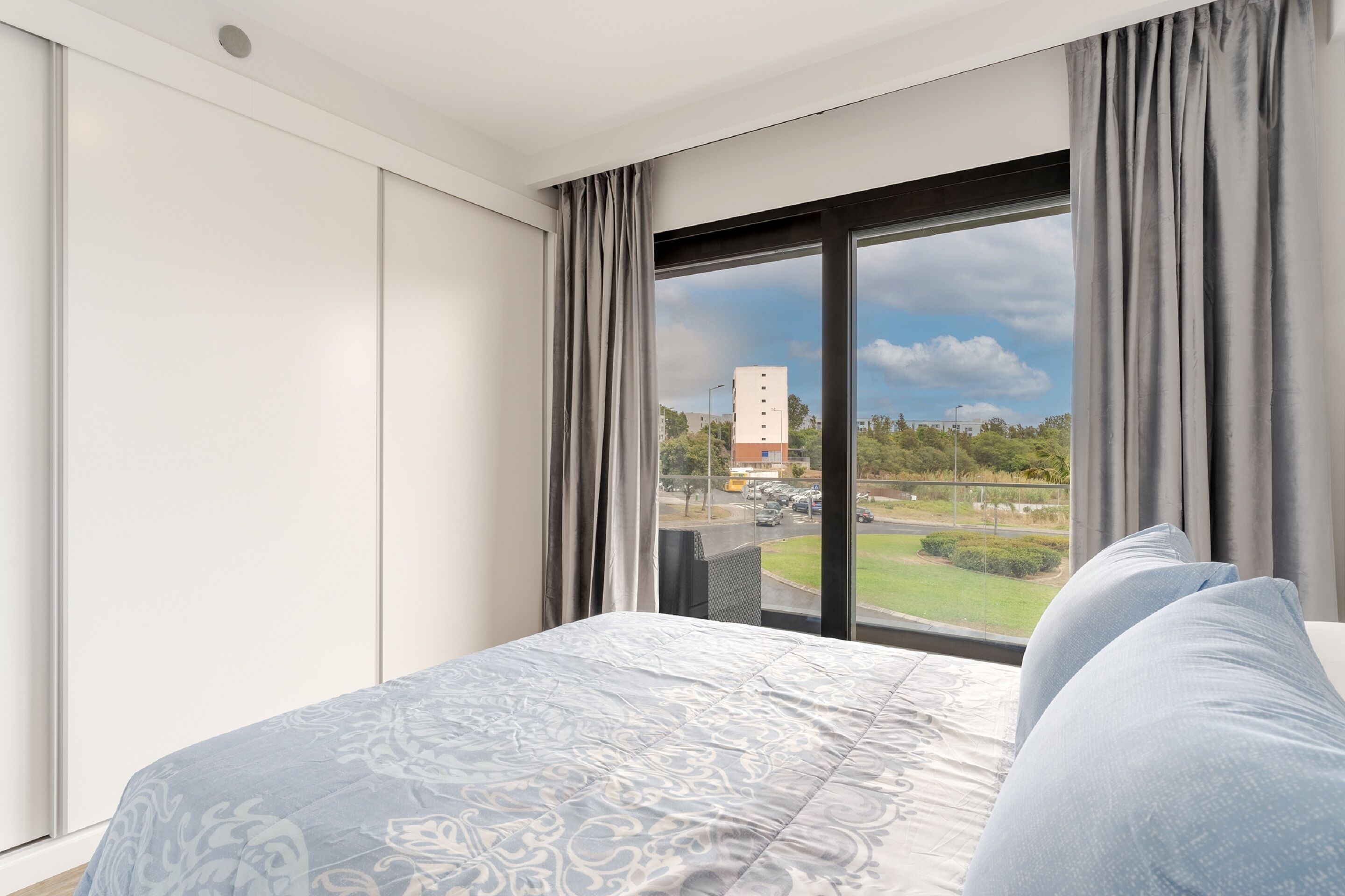 A modern hotel room with a large bed, a view of a parking lot and greenery outside, and a window with grey curtains.