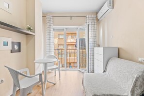 A bedroom with a bed, a small table, a chair, a dresser, and a balcony with a view of other buildings.