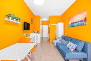 A compact living space with a kitchenette, a dining area, a sofa with patterned cushions, and a painting on the wall.