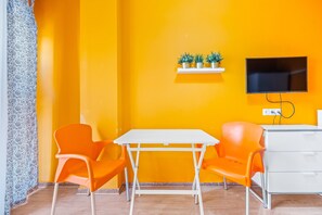 Una habitación con una mesa blanca, dos sillas anaranjadas, un aparador blanco, un televisor montado y una estantería con plantas en macetas.