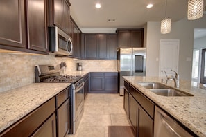 A modern kitchen with dark cabinetry, granite countertops, stainless steel appliances, and pendant lighting. - Affordable 3-Bedroom Townhouse With Backyard ~ Double Story (Pet Friendly!) (Rosenberg)