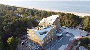 A modern building with a flat roof, surrounded by a forested area and a beach in the background.