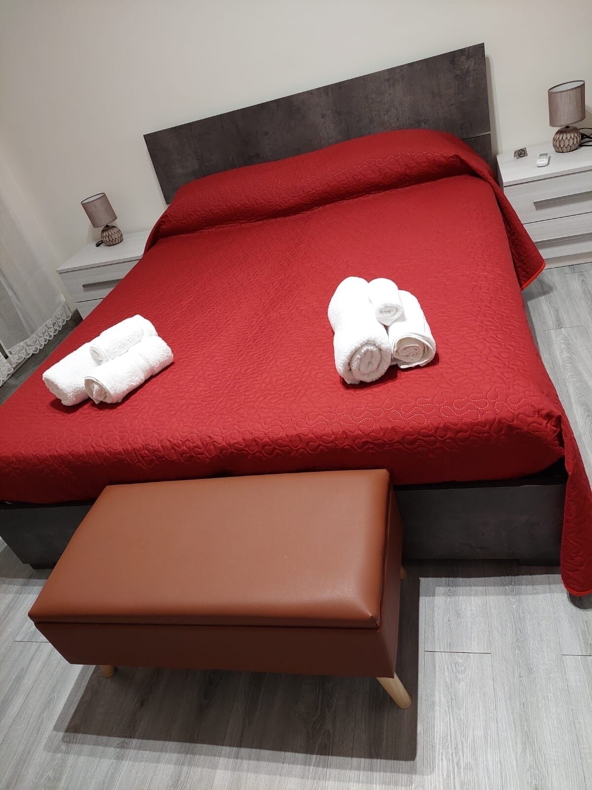 Una cama con ropa de cama roja, dos toallas blancas enrolladas, un banco marrón y una mesita de noche con lámpara.