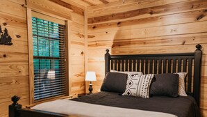 Une chambre à coucher de style cabane en bois, avec un lit, une table de chevet, une lampe et une fenêtre avec des stores.