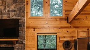 L’intérieur chaleureux d’un chalet avec une cheminée en pierre, des murs en bois, une table à manger et une grande fenêtre donnant sur la forêt.