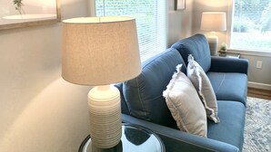 A living room with a blue sofa, a glass coffee table, and a lamp. There are two windows with blinds, a framed picture on the wall, and a rug on the floor.
