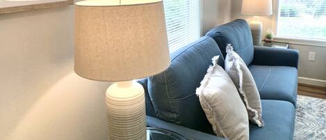 A living room with a blue sofa, a glass coffee table, and a lamp. There are two windows with blinds, a framed picture on the wall, and a rug on the floor.