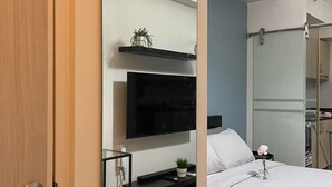 A modern hotel room with a large mirror, a flat-screen TV, a bedside table with a plant, a bed with white linens, and a shower area visible through the door.