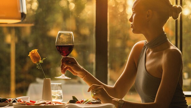 Una persona sentada a una mesa con una copa de vino tinto y una rosa, al aire libre con vistas a árboles y un atardecer.