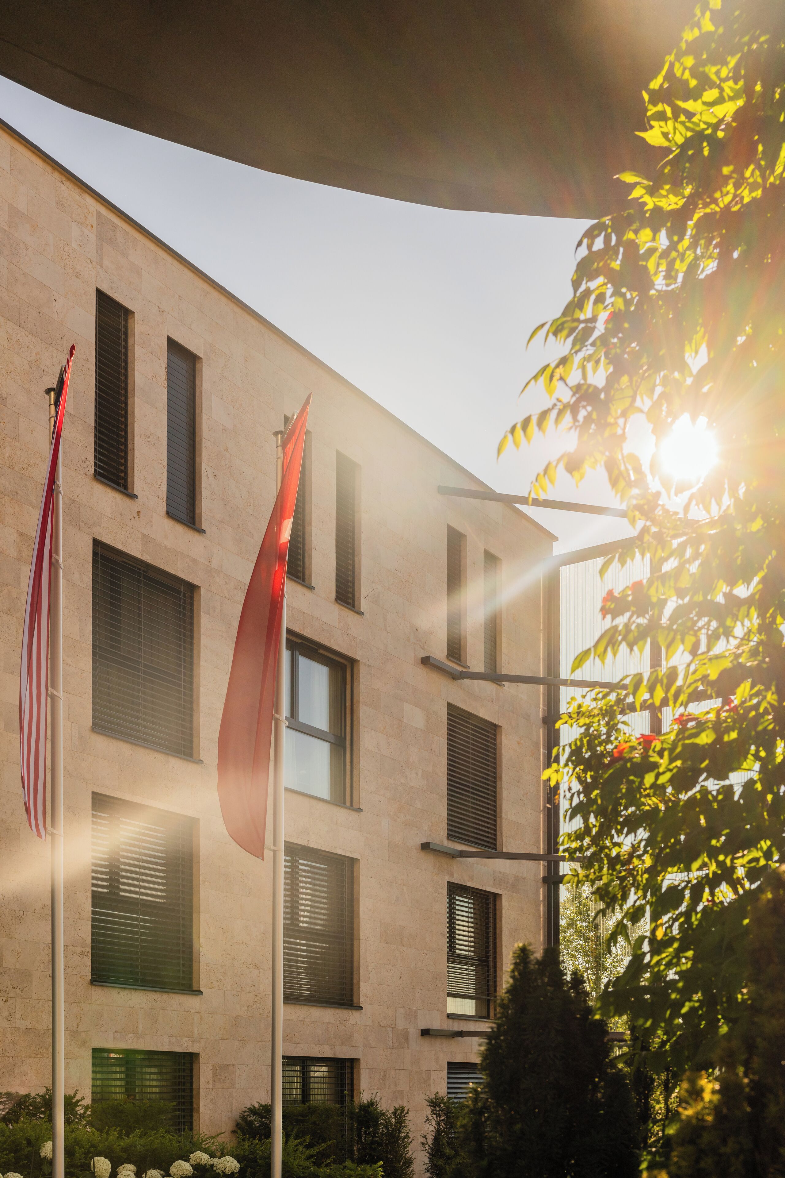 Un edificio con varias ventanas y banderas, incluyendo una bandera estadounidense, con la luz del sol filtrándose entre las hojas de un árbol.