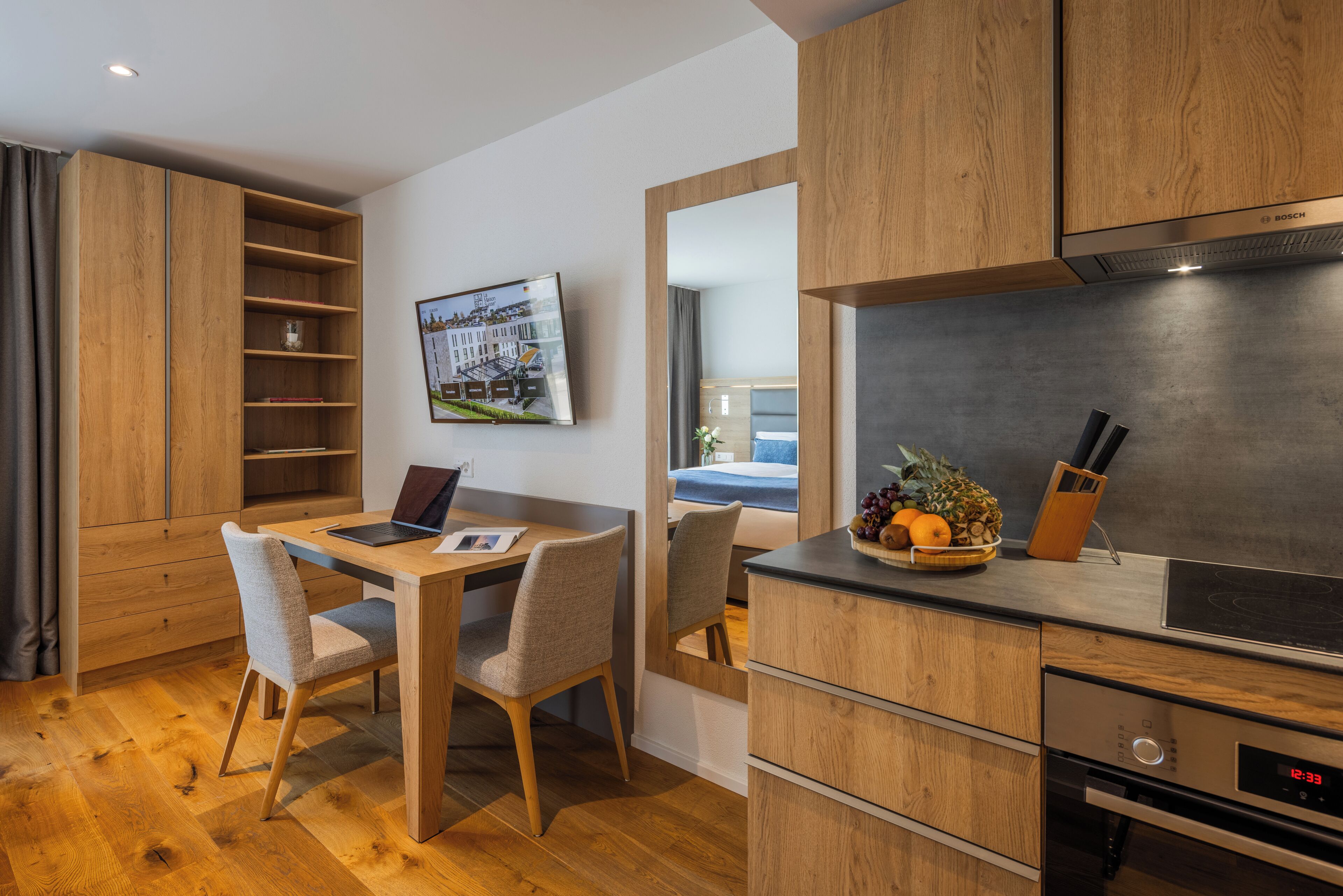 A modern kitchen with wooden cabinets, a built-in oven, and a digital clock displaying 12:33.