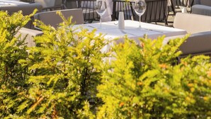 Un área de comedor al aire libre con mesas y sillas, rodeada de follaje y una cerca.