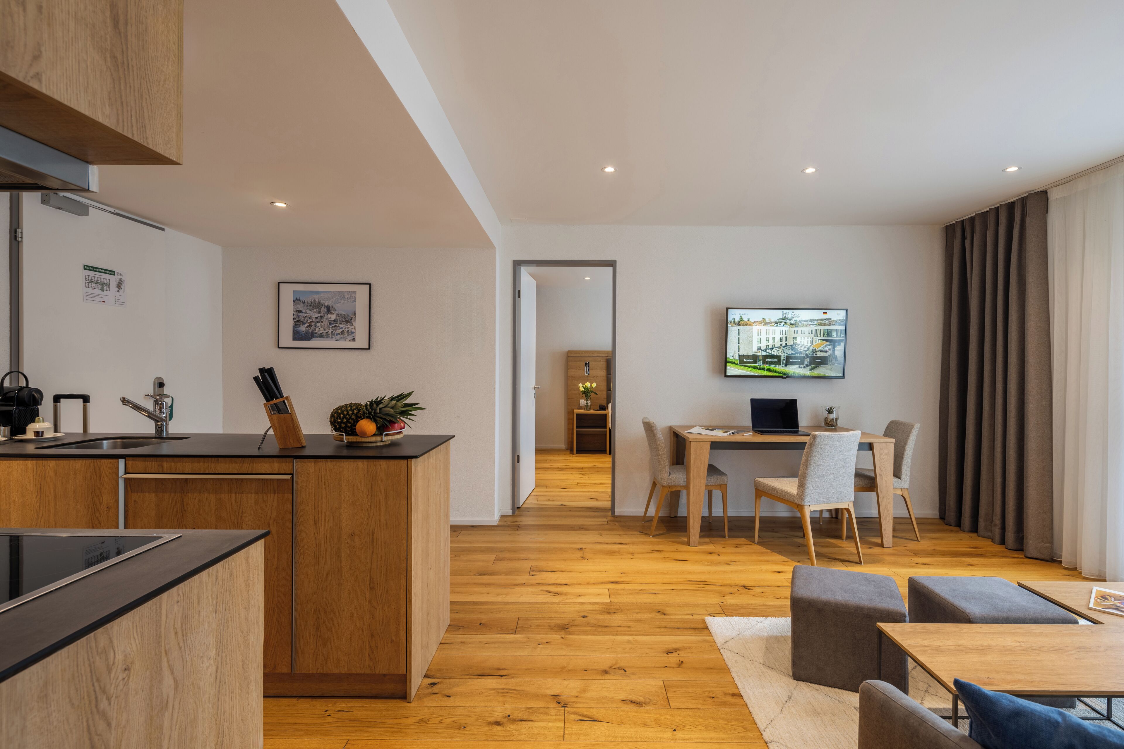 A modern kitchen with wooden cabinets and a central island, a dining area with a table and chairs, and a living space with a TV and seating.