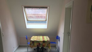 A small room with a tiled floor, a table with a colorful tablecloth, two blue chairs, a window with a view, and a door. - Pension Yildirim - quiet and central vacation apartment (1) (Leverkusen)