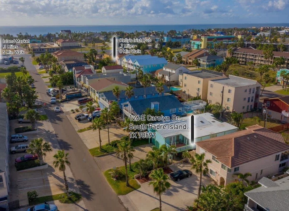 An aerial view of a coastal town with a prominent building at 126 E Carolyn Dr., South Padre Island, TX 78597. Nearby, Island Market is 3 miles away, and Kelly's Irish Pub is 7 miles away, both accessible by a 5-minute walk.