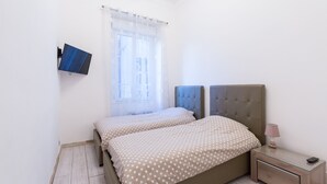 Ein Schlafzimmer mit zwei Betten, einem an der Wand befestigten Fernseher, einem Nachttisch mit Lampe und einem Fenster mit durchsichtigen Vorhängen.