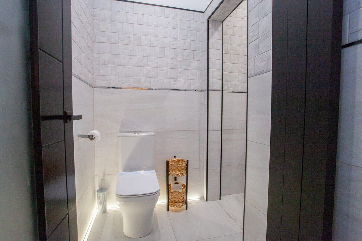 Uma casa de banho moderna com sanita branca, uma pequena cesta de vime e zona de duche revestida a azulejos.