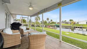 Un espace extérieur abrité, aménagé avec des meubles en osier, un barbecue et offrant une vue sur un canal où naviguent des bateaux.