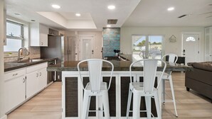 Une cuisine moderne avec des meubles blancs, un îlot central doté de tabourets de bar et une vue sur l’extérieur grâce à de grandes fenêtres.