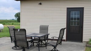 An outdoor patio with a table and four chairs, a door, and a view of a green field.
