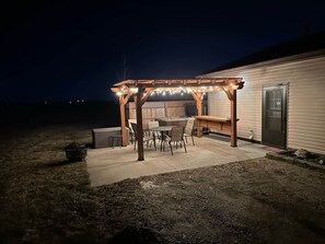 Un patio cubierto con una mesa, sillas, un banco de madera y una pérgola adornada con luces colgantes.