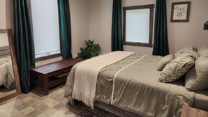 A bedroom with a bed, a bench, two windows with curtains, a plant, and a framed picture on the wall.