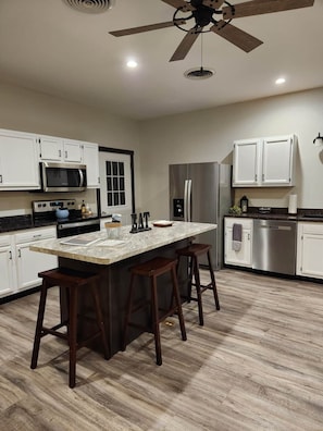 A modern kitchen with white cabinets, a central island with stools, stainless steel appliances, and a ceiling fan. - Prairieview Cottage Retreat - Hot Tub Sunsets (Sullivan)