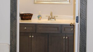 Une salle de bain avec un meuble-lavabo en bois foncé, un lavabo blanc, un miroir avec trois lampes et un panier posé sur le comptoir.