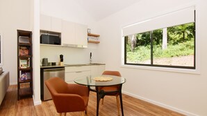 A compact kitchen with a dining area, a built-in bookshelf, and a large window with a view of greenery. - Rainforest River Studio Kangaroo Valley (Upper Kangaroo River)