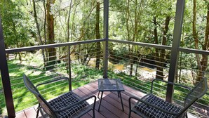 Une terrasse en bois avec des chaises et une table en métal, donnant sur une zone boisée traversée par un ruisseau.