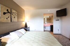 Una habitación de hotel con cama, una mesita de noche con lámpara, televisor montado en la pared y un baño visible a través de una puerta abierta.