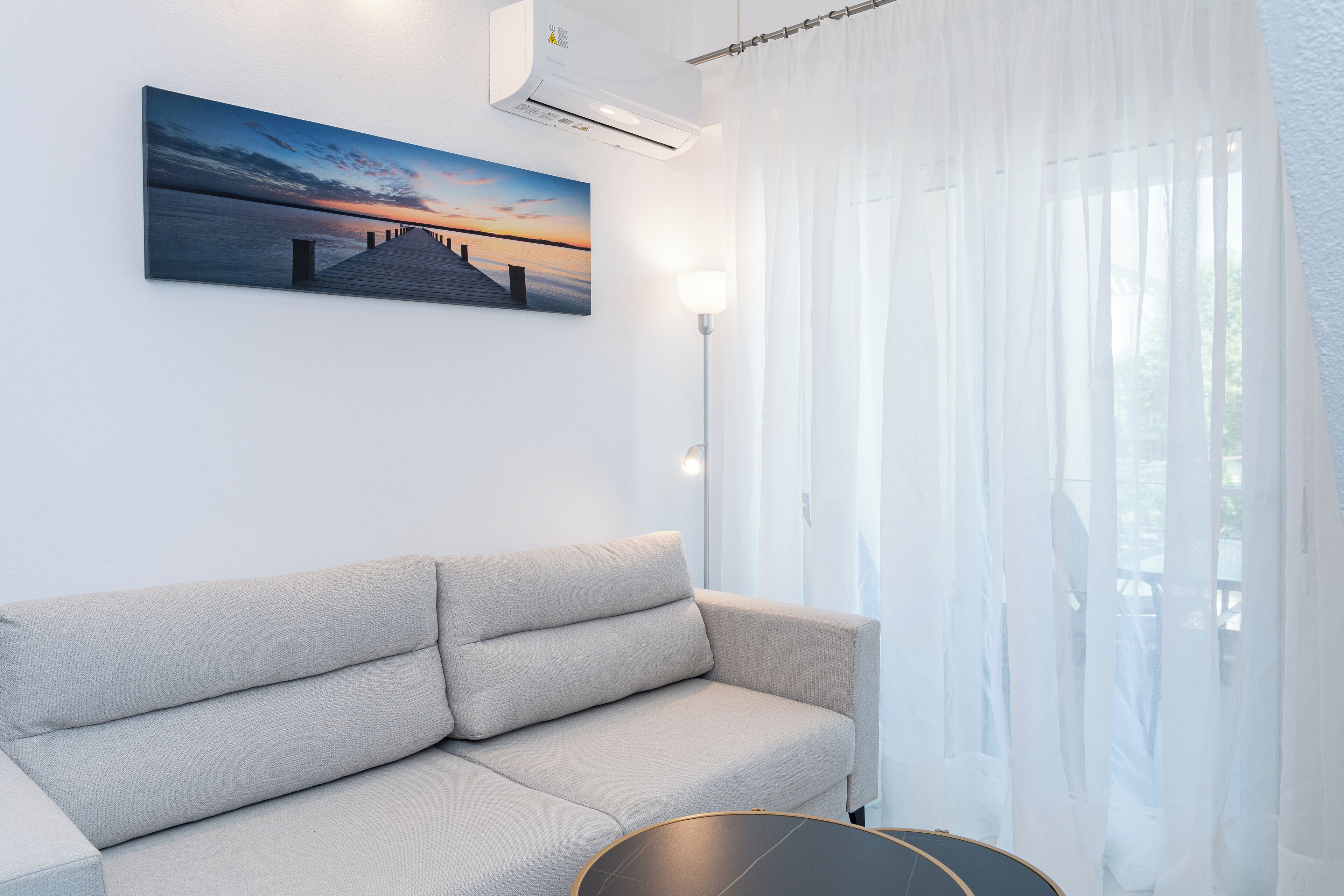 A modern living room with a beige sofa, a painting of a sunset over water, and a large window with sheer curtains.