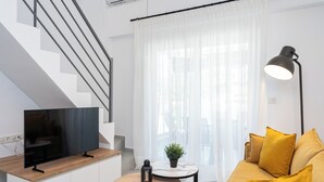 Un salon moderne avec un téléviseur à écran plat, une table ronde en bois, un lampadaire, un canapé avec des coussins jaunes et un escalier.