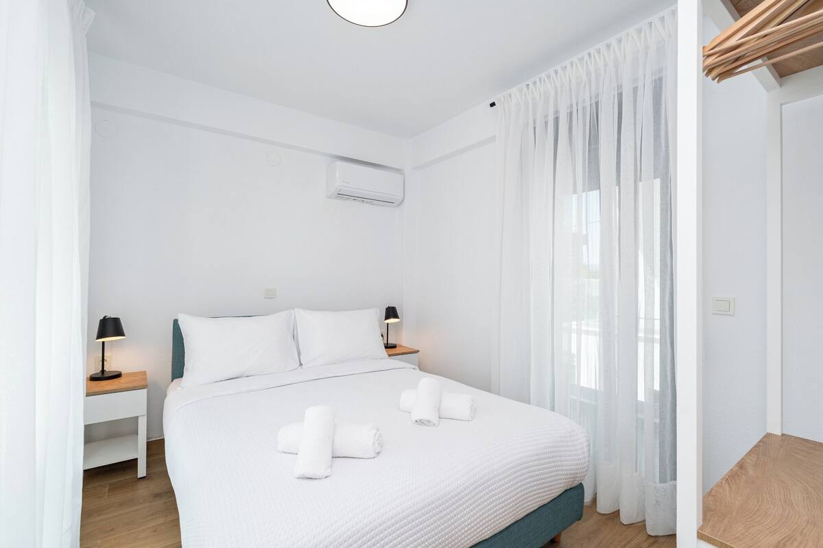 Ein Hotelzimmer mit einem Bett, zwei Nachttischen, einer Lampe, einem Fenster mit transparenten Vorhängen und einer Klimaanlage.