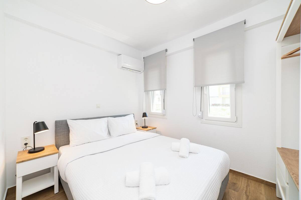 Ein schlicht eingerichtetes Hotelzimmer mit einem Bett, Nachttischen, einem Schreibtisch und einem Fenster mit Jalousien.