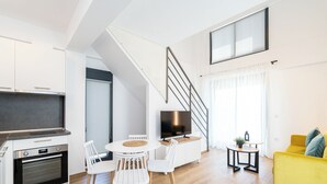 Une cuisine moderne dotée d’un four encastré, un coin repas avec une table ronde et des chaises blanches, un fauteuil jaune et un escalier menant à un niveau supérieur.