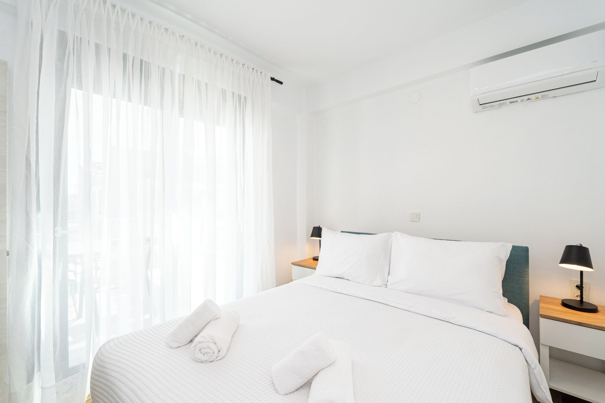 A hotel room with a neatly made bed, a bedside table with a lamp, and a window with sheer curtains.
