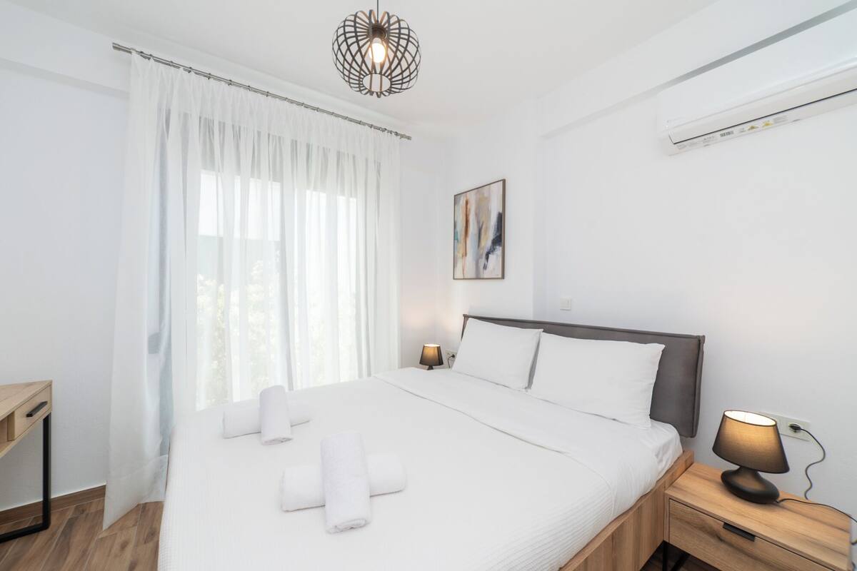 Ein ordentlich bezogenes Bett mit weißen Laken, eine Nachttischlampe und ein an der Wand befestigtes abstraktes Gemälde.