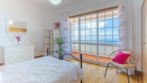 Ett sovrum med en säng, en stol med en rosa kudde, en byrå, en spegel och ett fönster med utsikt över havet.