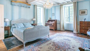 A bedroom with a four-poster bed, a chandelier, a wooden dresser, and a large window with curtains.