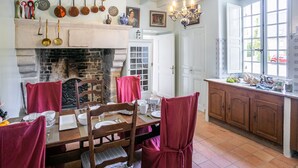 A dining area with a fireplace, wooden chairs, a table set for four, and a kitchen area with cabinets and a window.