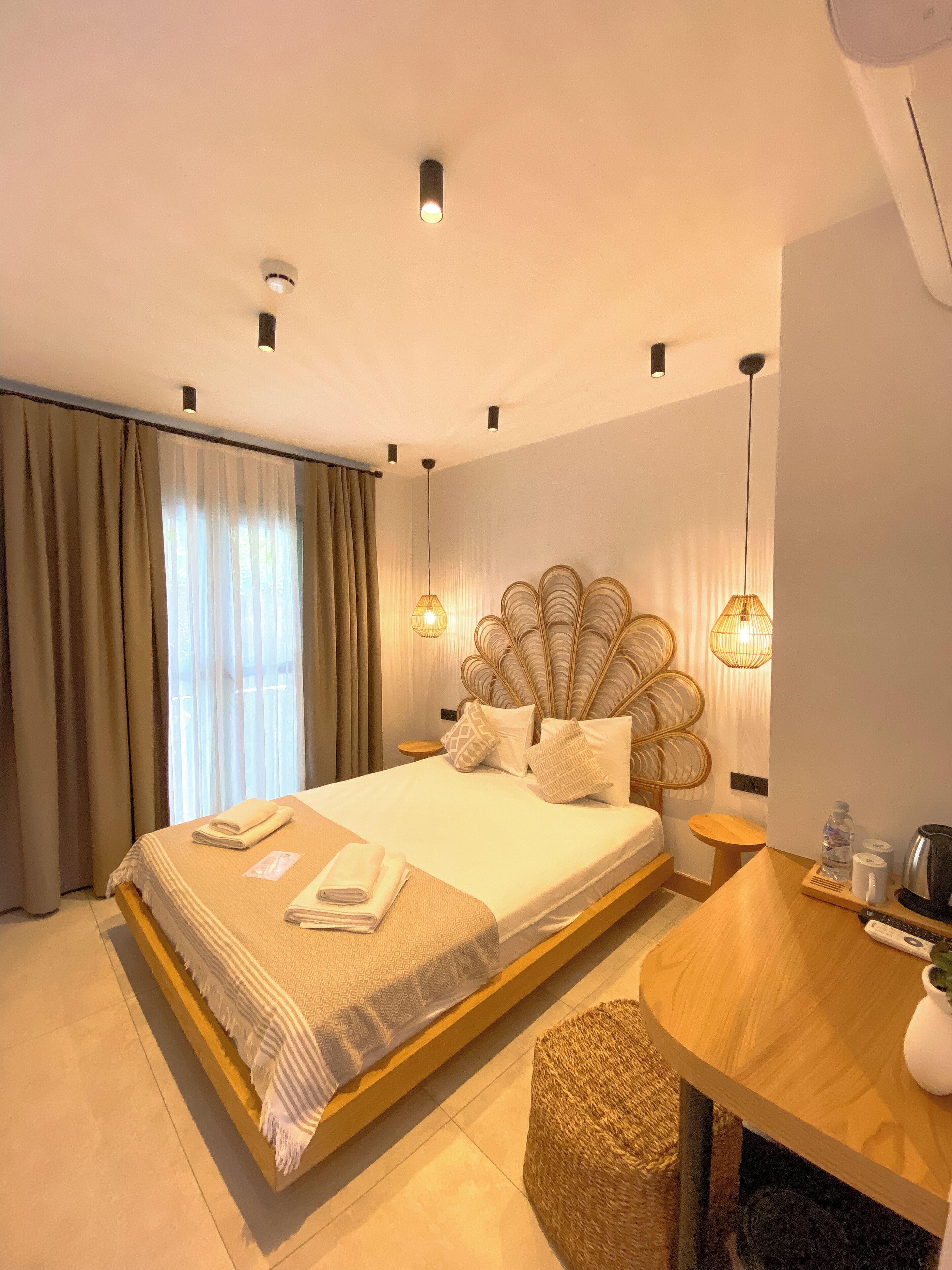 A modern hotel room with a large bed, a wooden side table, a small round table, and a woven stool.